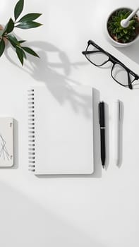 A neat workspace features a spiral notebook, black and white pens, and glasses next to a small potted plant. Sunlight casts gentle shadows on a clean surface, creating a calm atmosphere.