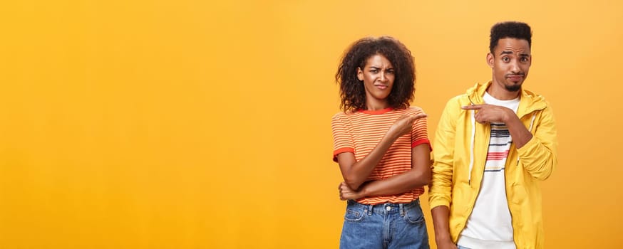 Friends getting shocked someone thought they are dating. Displeased mocking african american female frowning and grimacing from aversion while pointing at guy who indicating at girl over orange wall.