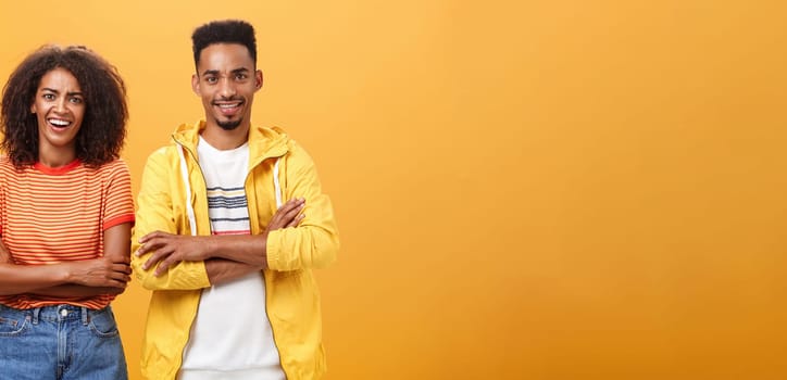 Portrait of surprised sarcastic african american woman with afro hairstyle standing with cute boyfriend crossing arms on chest laughing from scorn and fun over orange background. Copy space
