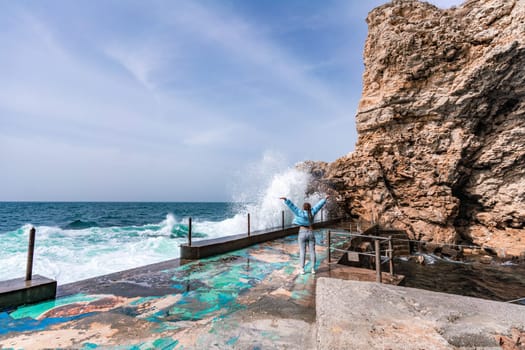 A woman in a blue jacket stands on a rock above a cliff above the sea and looks at the raging ocean. Girl traveler rests, thinks, dreams, enjoys nature. Peace and calm landscape, windy weather