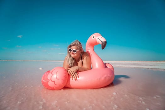 A woman in sunglasses lays on a pink flamingo inflatable in a shallow body of water with a pink tint. The sky is blue.