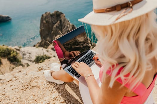 A woman is sitting on a rock by the ocean, typing on her laptop. Concept of relaxation and leisure, as the woman enjoys the beautiful view of the water while working on her computer