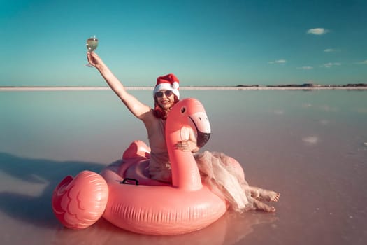 A woman wearing a Santa hat sits on a pink flamingo float in a body of water. He is smiling. There are white posts in the background.