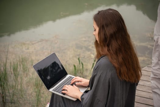 A woman is sitting on a dock with a laptop open in front of her. She is typing on the keyboard and she is focused on her work. The scene suggests a sense of solitude and concentration