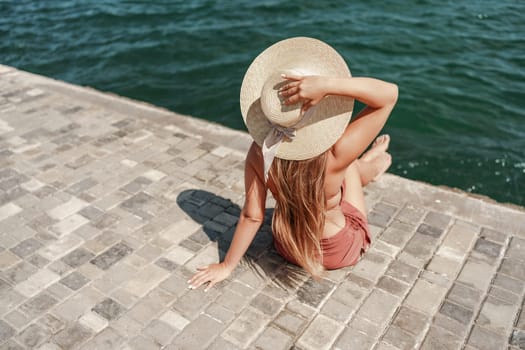 A woman in a swimsuit sits with her back holding a hat, looks at the ocean, sunny day, relaxes