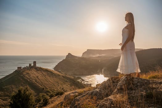 A woman stands on a rocky hill overlooking the ocean. She is wearing a white dress and she is enjoying the view. The scene is serene and peaceful, with the sun shining brightly in the background