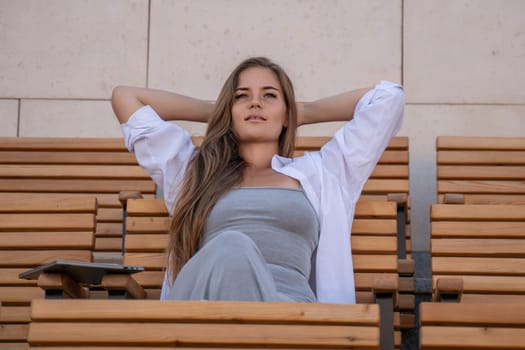 A woman with long brown hair sits on a wooden bench, wearing a white shirt and a gray dress, her hands behind her head.