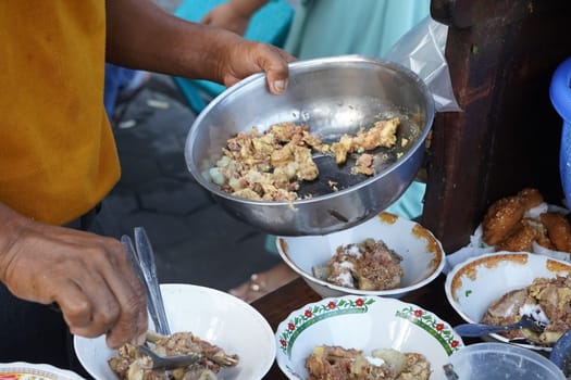 A street food vendor in Surabaya, Indonesia, is preparing a hearty soup with beef ribs and other ingredients. He's using a large metal bowl to mix the ingredients before serving them in smaller bowls.