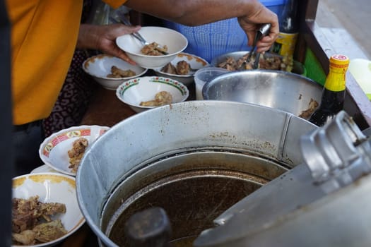 A street food vendor in Surabaya, Indonesia, is adding cooked beef ribs to bowls of soup. The vendor is using tongs to carefully place the ribs into the bowls, which are decorated with colorful patterns.