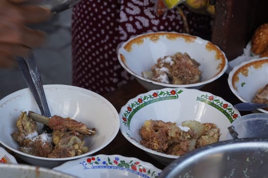 A street food vendor in Surabaya, Indonesia, is adding cooked beef ribs to bowls of soup. The vendor is using tongs to carefully place the ribs into the bowls, which are decorated with colorful patterns.