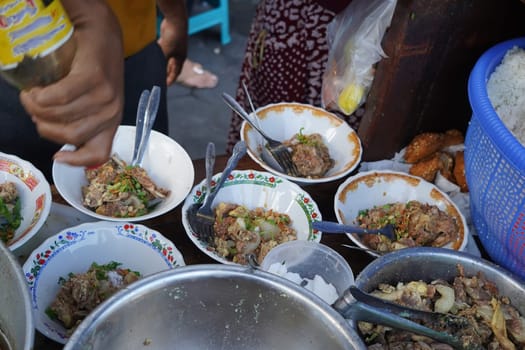 A street food vendor in Surabaya, Indonesia, is adding cooked beef ribs to bowls of soup. The vendor is using tongs to carefully place the ribs into the bowls, which are decorated with colorful patterns.