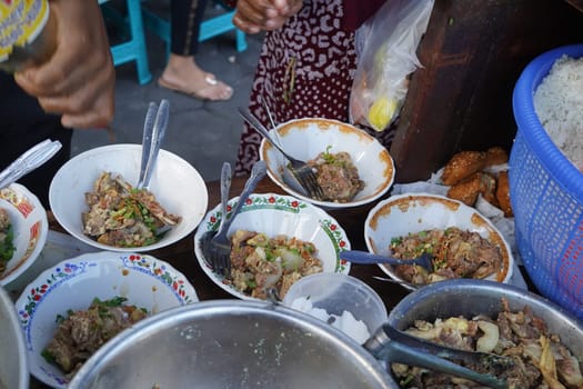 A street food vendor in Surabaya, Indonesia, is adding cooked beef ribs to bowls of soup. The vendor is using tongs to carefully place the ribs into the bowls, which are decorated with colorful patterns.