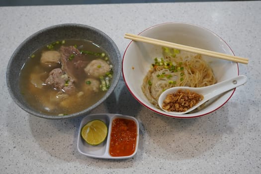 A bowl of savory bakso soup with meatballs, noodles, and broth, served with a small plate of lime wedges and chili sauce, accompanied by a bowl of vermicelli noodles and a small plate of fried shallots.