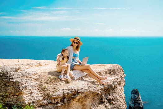 A woman and a child are sitting on a rock overlooking the ocean. The woman is using a laptop while the child looks on. Concept of relaxation and bonding between the two