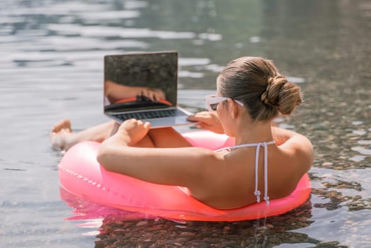 A woman is sitting on a pink inflatable raft in the water, using a laptop. Concept of relaxation and leisure, as the woman is enjoying her time in the water while working on her laptop
