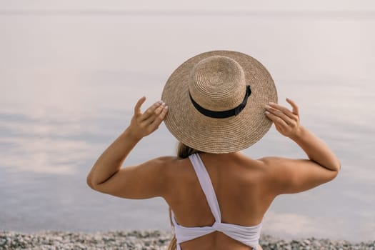 A woman wearing a straw hat is standing on a beach. She is holding her hat up to her face