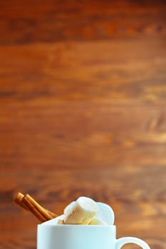 Close-up of a warm drink topped with marshmallows and cinnamon sticks, set against a wooden background.