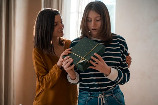 Two happy young girls laugh, friends hug and give each other gifts during the holiday. Women surprise and congratulate on the anniversary of their relationship in their cozy home.