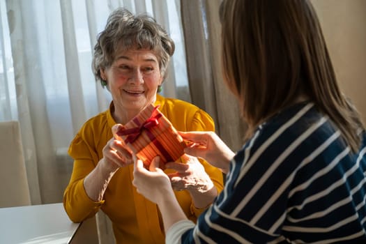 A young girl congratulates an elderly woman, her mother or grandmother, daughter holds in her hands a small cute beautifully wrapped gift for her grandparent, gives a surprise with love and care.