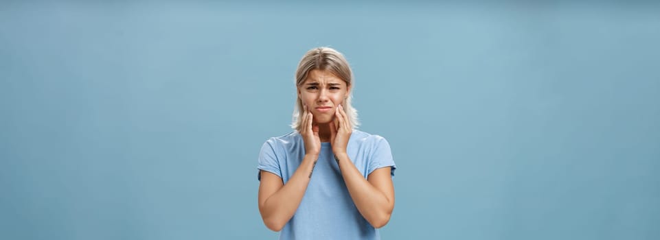Girl feeling pain in teeth after drinking hot coffee. Displeased unhappy troubled young girl with blond hair frowning feeling discomfort in mouth holding hands on cheeks suffering toothache.