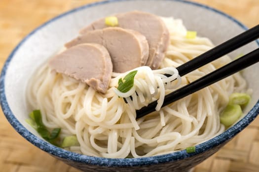A close up photo of using chopsticks  to eat a bowl of noodles topped with pork slices and green onions.