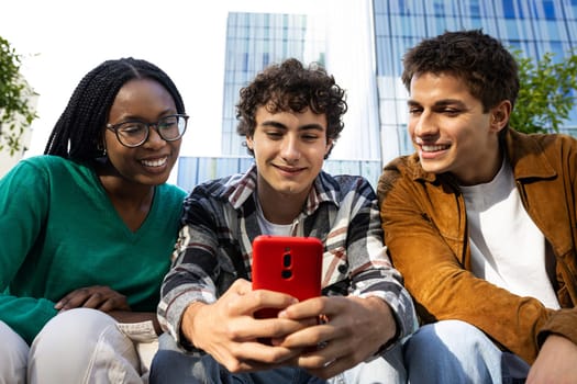 Happy college students looking at mobile phone laughing together. Multiracial teenagers using smartphone outdoors. Youth lifestyle and social media addiction concept.