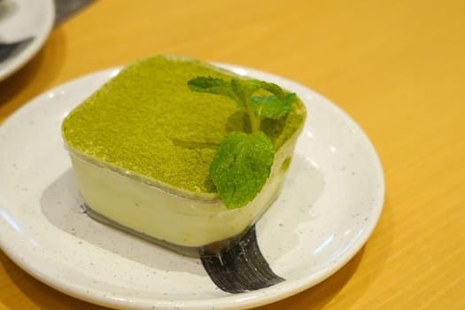 A square matcha cake topped with a mint leaf, served on a white plate with a black brushstroke design.