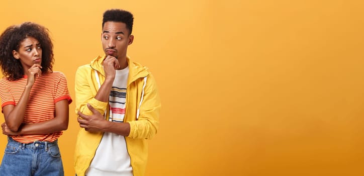 Silly confused girl standing gloomy and worried pursing lips and frowning holding hand on chin looking at boyfriend confused while guy glancing at girlfriend with curious expression over orange wall. Lifestyle.