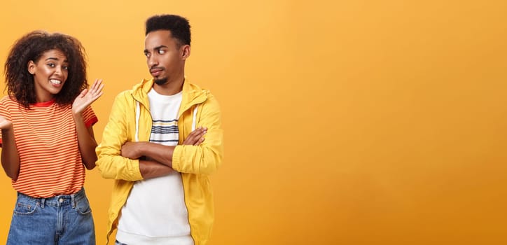 Guy thinks his friend weirdo making dumb thinks looking at cute girl with suspicious look crossing arms on chest raising eyebrow questioned while girlfriend saying sorry shrugging over orange wall. Lifestyle.