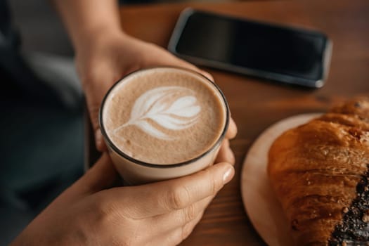 A woman holding a coffee cup with a leaf design on it. She is sitting at a table with a croissant and a cell phone