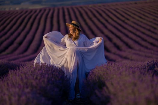 Woman poses in lavender field at sunset. Happy woman in white dress holds lavender bouquet. Aromatherapy concept, lavender oil, photo session in lavender.