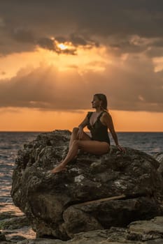 A woman in a swimsuit sits on a rock by the ocean, gazing at the sunset. Golden light from the sun pierces through the clouds.