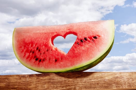 Ripe watermelon slice with heart shaped hole on blue background of the blue sky with little cloud on a hot summer day