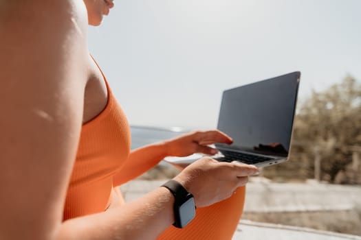 A woman wearing orange sportswear sits outdoors and uses a laptop. The screen is dark and the background is blurred.