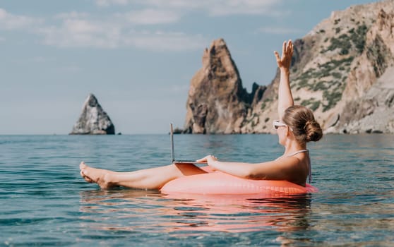 Woman freelancer works on laptop swimming in sea on pink inflatable ring. Pretty lady typing on computer while floating in the sea on inflatable donut at sunset. Freelance, remote work on vacation