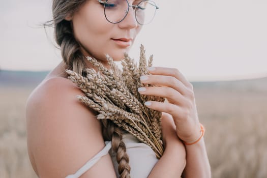 A woman is in a wheat field holding a bunch of wheat close to her body, her arms crossed, wearing a white top and glasses, she is looking down at the wheat.