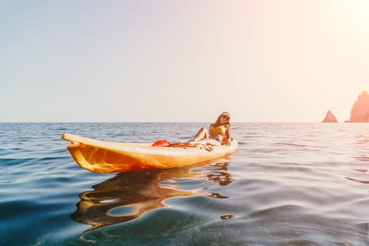 A woman kayaks on a calm sea with rocky cliffs in the distance. The sun is shining and the sky is blue.