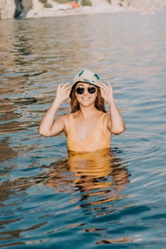 A woman in a yellow swimsuit and sunglasses stands in clear blue water, adjusting her bucket hat.