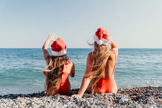 Two women in red swimsuits and Santa hats sit on a pebbled beach with the ocean in the background.