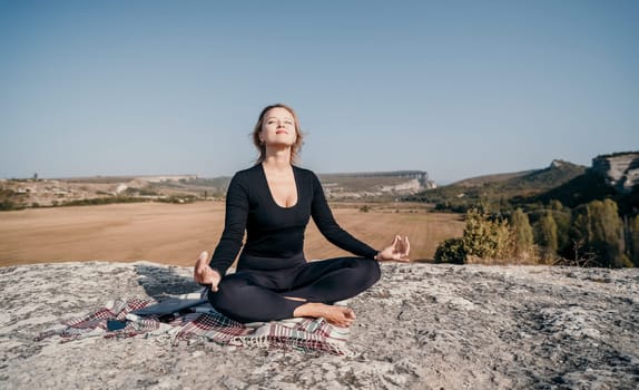 A woman in black clothing sits in a meditative pose on a rocky mountaintop, enjoying the serenity of the surrounding landscape.