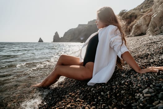 A woman wearing a white shirt and a black swimsuit sits on a pebbly beach, her feet in the water. The sea and distant rocky cliffs are in the background.