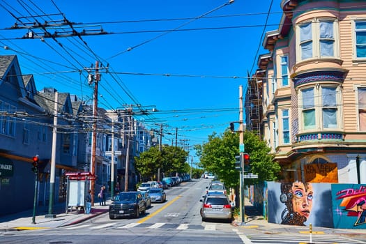 Image of Street with cars going down hill and yellow house with bay windows and concrete wall covered in art