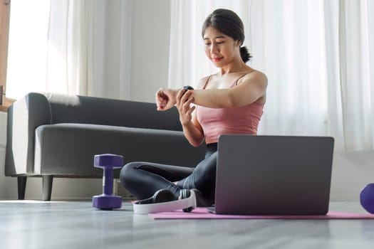 A young woman sits and looks at a smartwatch while exercising in front of her laptop. Wear a sports bar outfit. Do yoga and lift dumbbells on the exercise mat..
