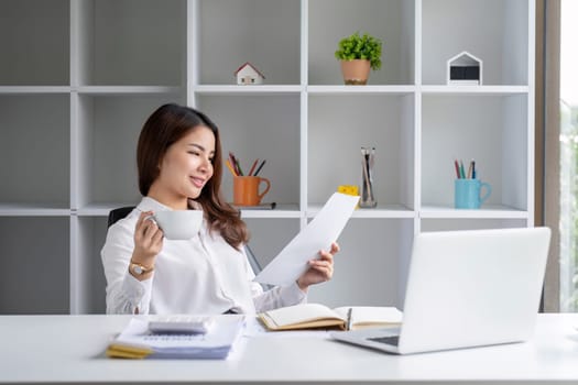 Young office worker sitting at table drinking coffee while checking documents and working on laptop..