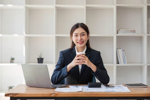 Photo of positive Asian businesswoman holds a cup of coffee imagines plan holiday in office..