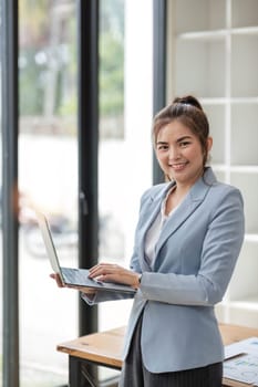 Portrait, Professional and confident Asian businesswoman or female executive manager in formal suit and use laptop. standing, leaning on table, holding laptop and using laptop computer..