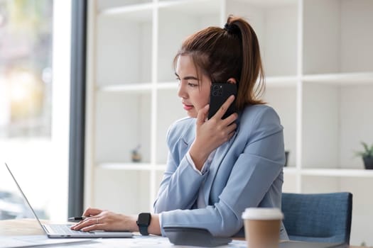 Attractive Asian woman using laptop in online meeting. Businesswoman using mobile phone talking with colleague sitting at table in modern office.