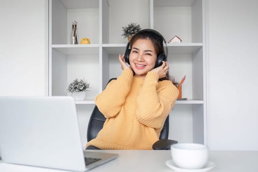 Young woman wearing headphones using laptop sits on sofa in living room, listening to music and drinking coffee, relaxing..