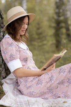 A young beautiful woman in a dress and a round hat reads a book outdoors in the forest and drinks tea. Romantic and vintage photo of a beautiful girl. Reading and relaxation