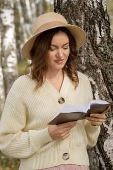 A young beautiful woman in a dress and hat stands and reads a Bible book in nature. Romantic and vintage photo of a beautiful girl. The book has the Bible written on it. Faith in God concept